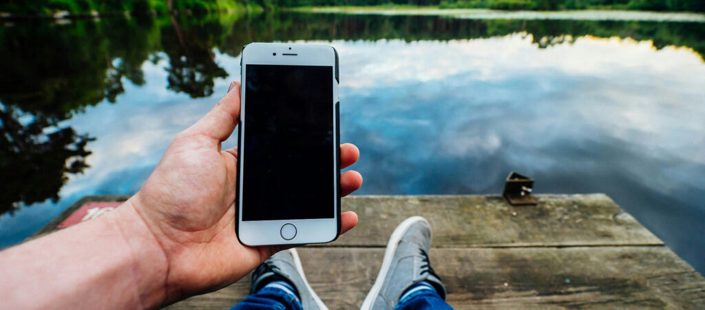 Person holding up a cell phone while sitting on a dock overlooking a lake