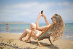 A woman in a white bikini sitting on a beach chair in the sand while she uses a smartphone.